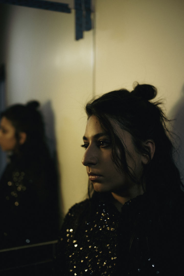 In a warmly lit, white-walled room, a woman in black sequins stares into the distance. Behind her, a mirror reflects her back.